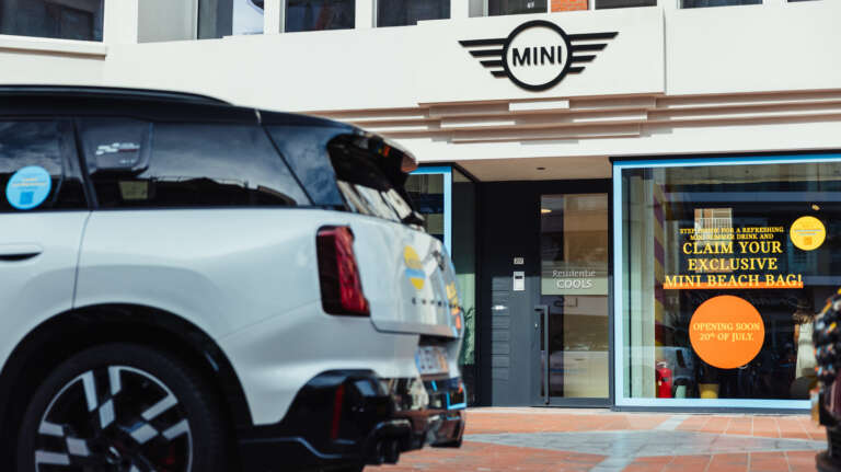 Outside view of a MINI dealership with a parked car and a window promoting an exclusive beach bag.