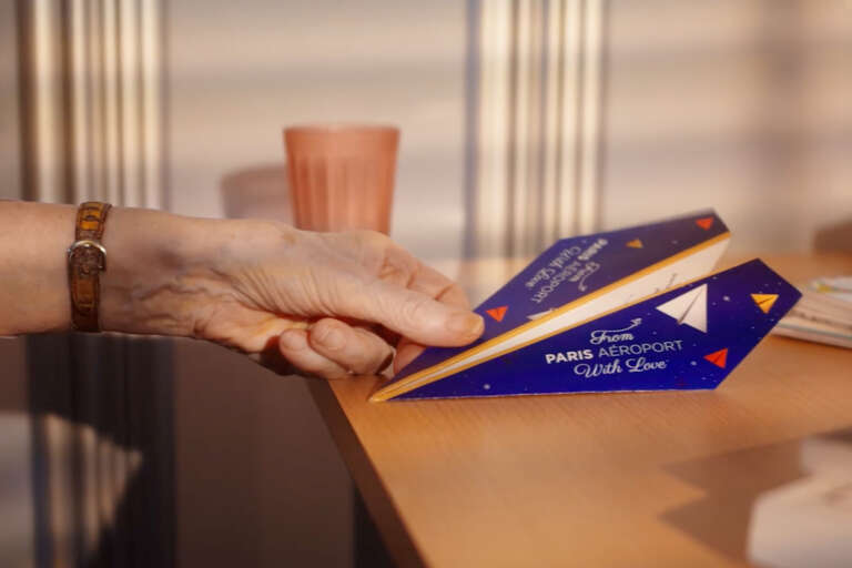 Une main tient un avion en papier bleu avec le message « From Paris Aéroport With Love »