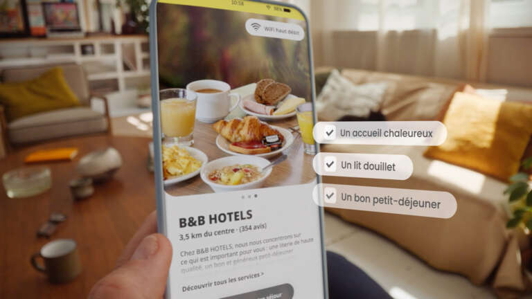Hand holding a smartphone displaying a B&B HOTELS page with a delicious breakfast and the criteria: “A warm welcome,” “A cozy bed,” “A good breakfast.”