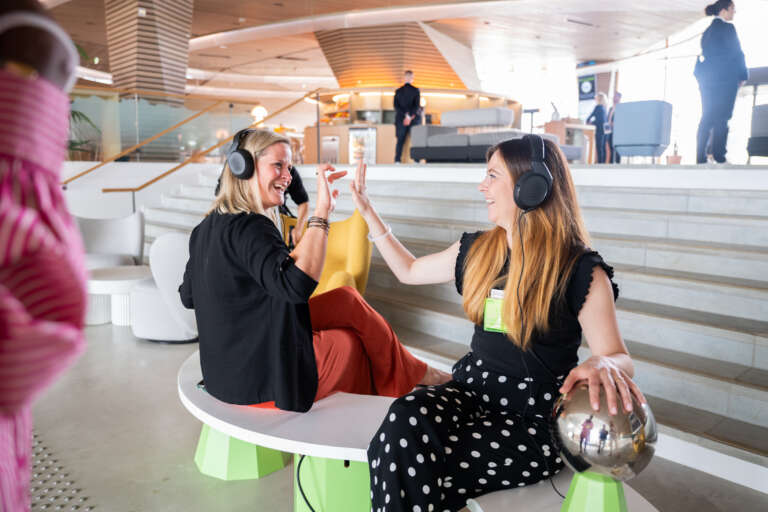 Deux participantes souriantes portant des casques audio interagissent dans un espace d'accueil moderne et lumineux.