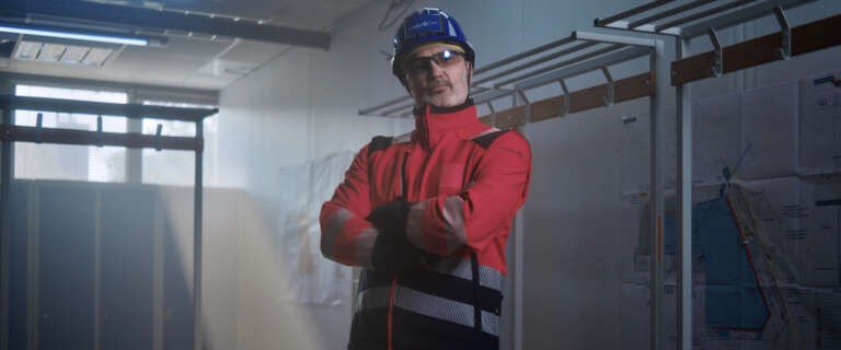 Un ouvrier en tenue de sécurité rouge et bleue, casque sur la tête et lunettes de protection, pose fièrement dans un vestiaire de chantier.