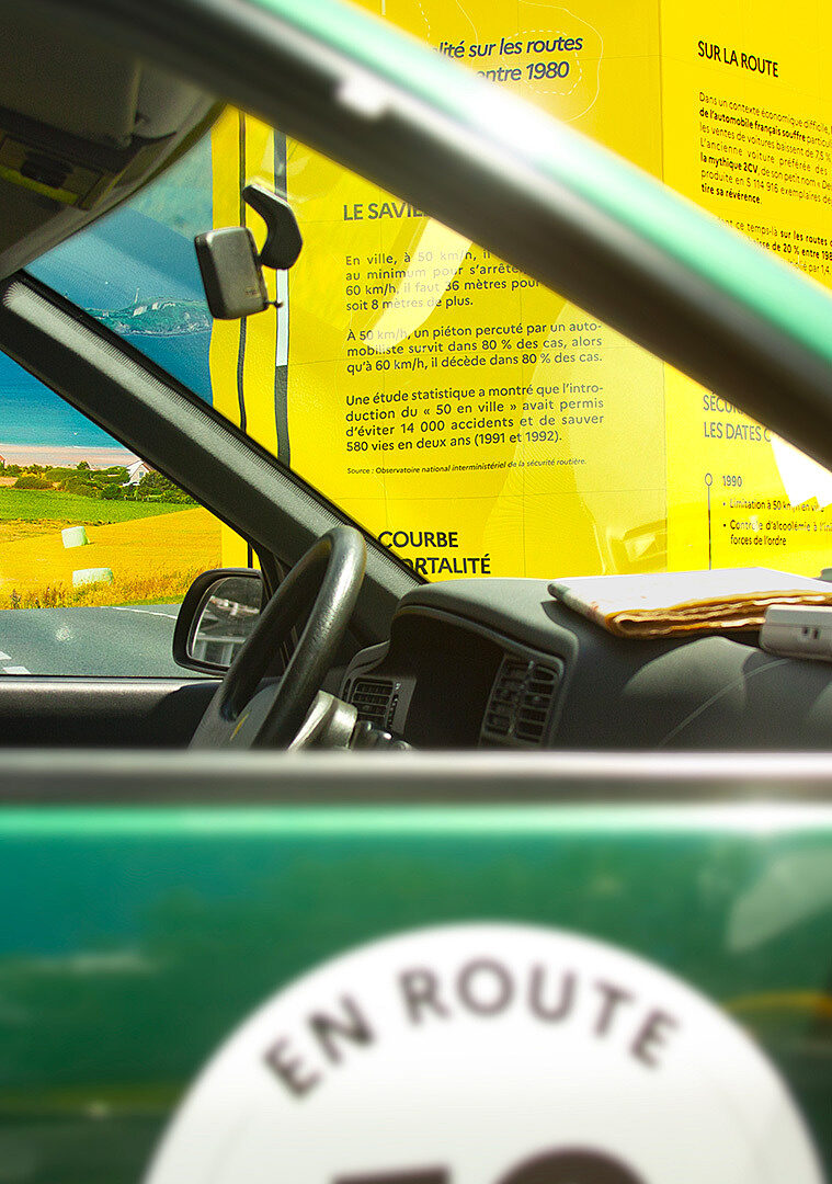 Vue intérieure d’une voiture ancienne avec en arrière-plan un panneau jaune retraçant l’histoire de la sécurité routière.