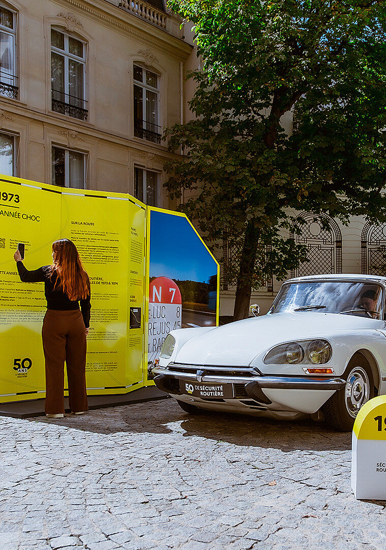 Une femme interagit avec une exposition sur la sécurité routière aux côtés d’une Citroën DS blanche.