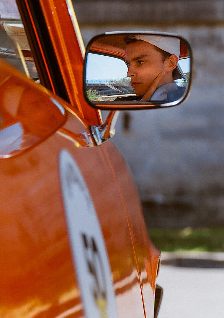Un jeune homme vu à travers le rétroviseur d’une voiture orange vintage.