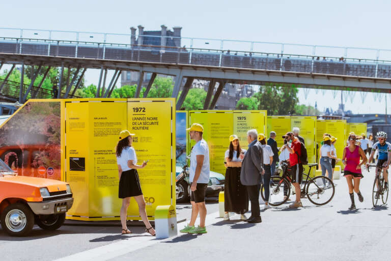 Vue d’ensemble de l’exposition itinérante avec panneaux jaunes et voitures anciennes, en bord de Seine.