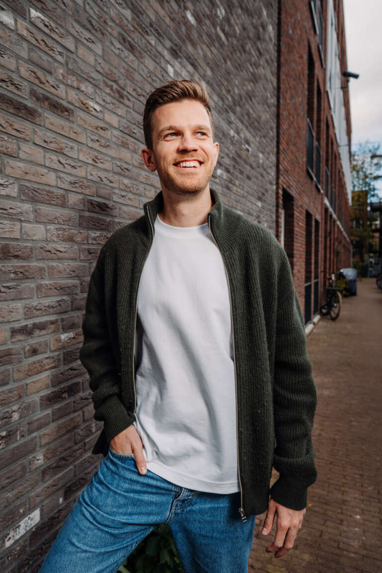 Portrait d’un jeune homme souriant en extérieur, adossé à un mur de briques.