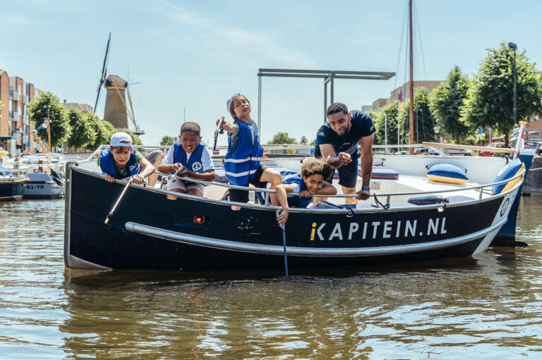Des enfants apprennent à naviguer sur un bateau avec un accompagnateur, dans un cadre urbain avec un moulin à vent en arrière-plan.