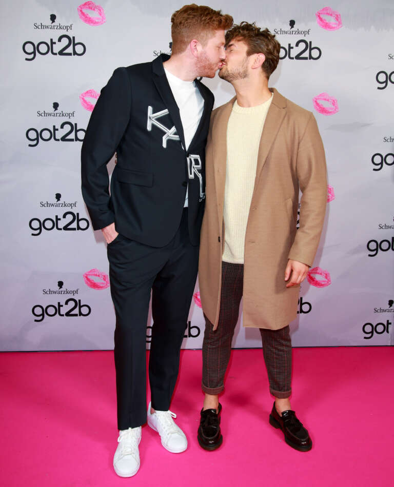 Two men sharing a kiss on the pink carpet, celebrating self-expression at the got2b event.